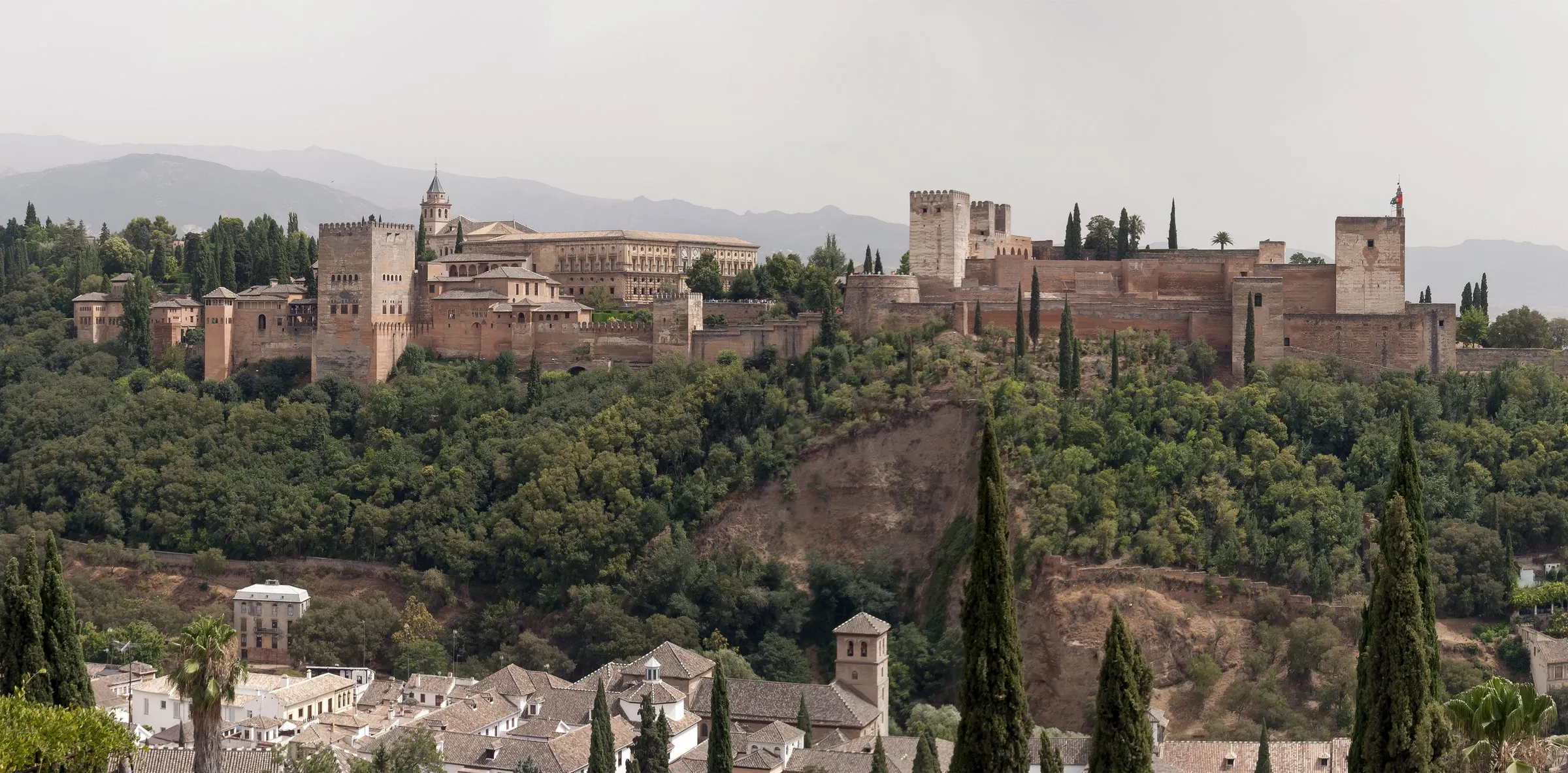 Die Alhambra mit Palästen und Türmen auf einem bewaldeten Hügel über den Dächern von Granada, dahinter Berge im Dunst